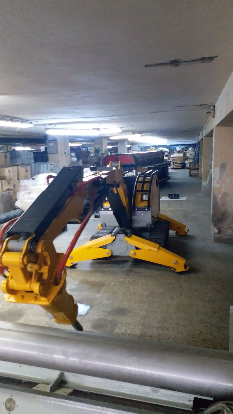 A yellow and black DEMOLISHING UNIT BROKK 180 REV-F1 stands in a dim basement, surrounded by boxes, shelves, and concrete pillars. The worn floor and damaged ceiling emphasize this powerful industrial robotic arm.