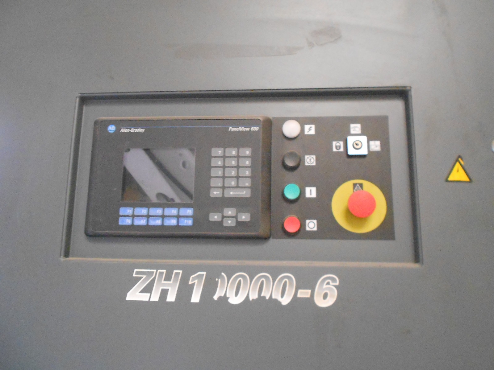 Close-up of an industrial control panel for the ATLAS COPCO ZH 10000-6-7PL AIR COMPRESSOR, with a screen, keypad, buttons, emergency stop, and the label ZH1000-6 below. Grey background shows warning symbols.