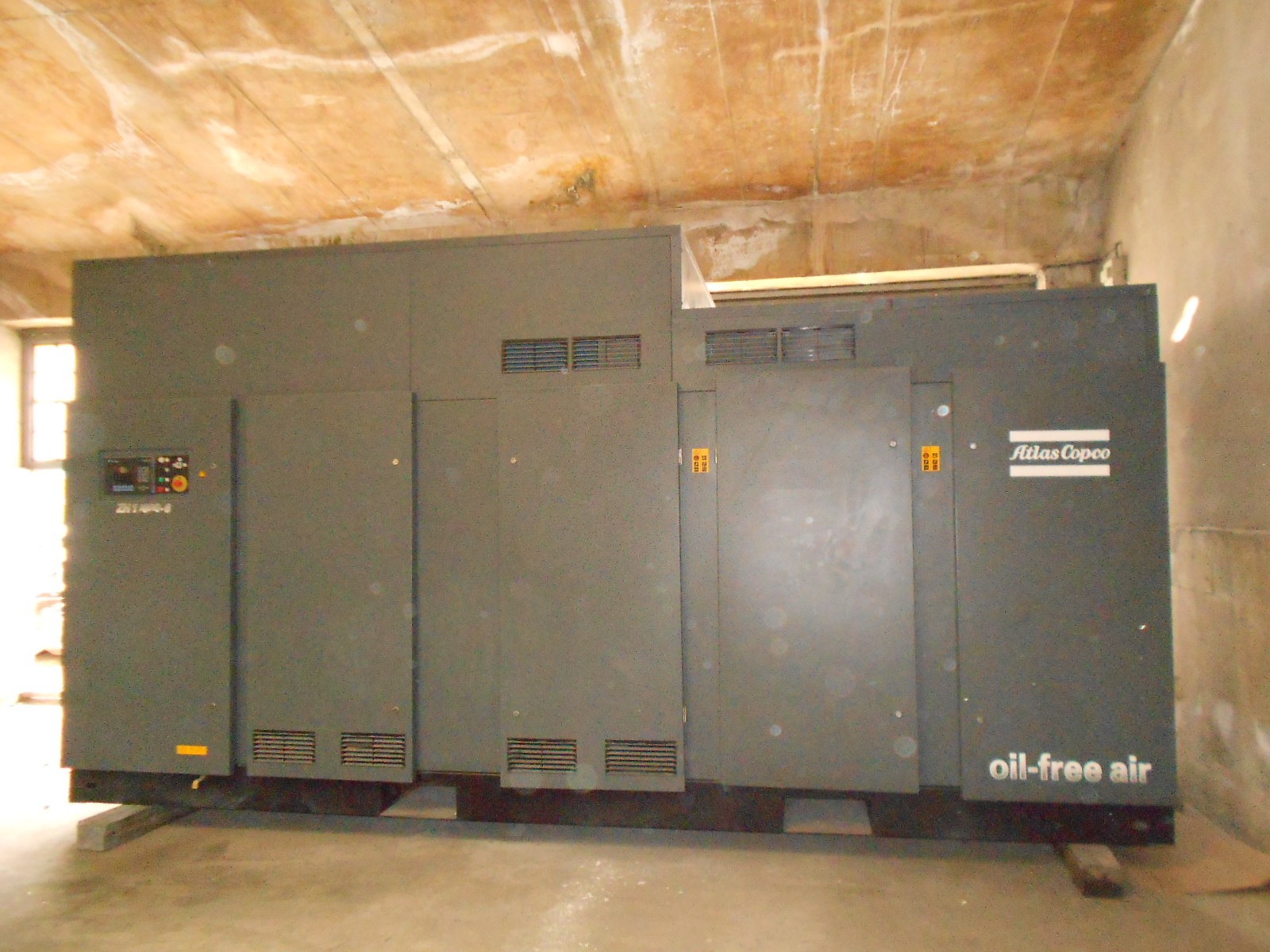 The ATLAS COPCO ZH 10000-6-7PL AIR COMPRESSOR, a large industrial oil-free unit, is installed in a concrete room with a worn ceiling, equipped with control panels and ventilation grilles.