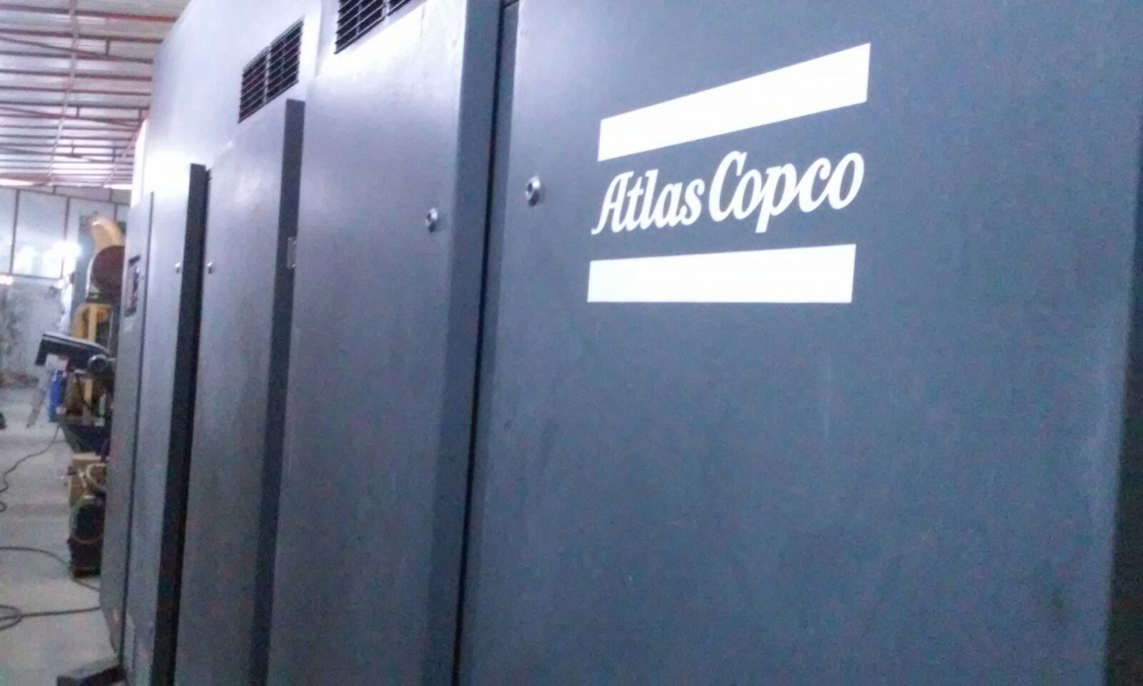 An ATLAS COPCO ZH 10000-6-7PL AIR COMPRESSOR with the white logo is installed indoors, likely in a factory or workshop, with other equipment and pipes visible in the background.