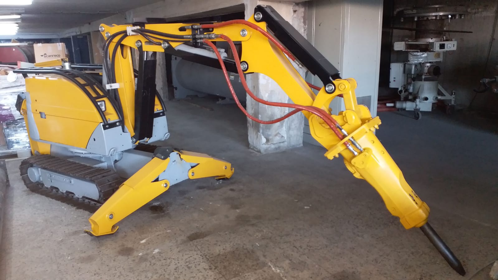 A yellow and gray DEMOLISHING UNIT BROKK 180 REV-F1 with caterpillar tracks and a large hydraulic breaker attachment is positioned indoors on a concrete floor.