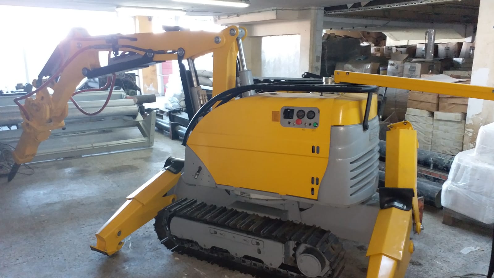A compact yellow and gray DEMOLISHING UNIT BROKK 180 REV-F1 tracked robotic excavator is parked indoors, surrounded by boxes, shelves, and industrial equipment with its articulated arm extended to the side.