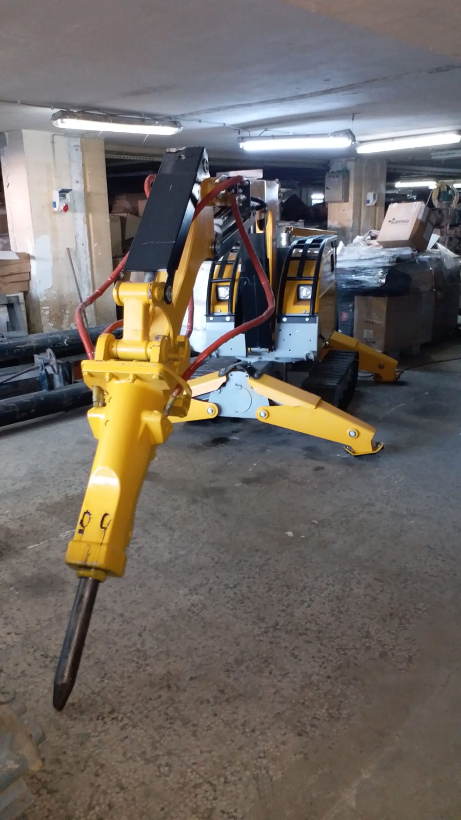 A DEMOLISHING UNIT BROKK 180 REV-F1 with an extended hydraulic arm and breaker is stationed in a cluttered indoor workspace with concrete floors and assorted equipment in the background.