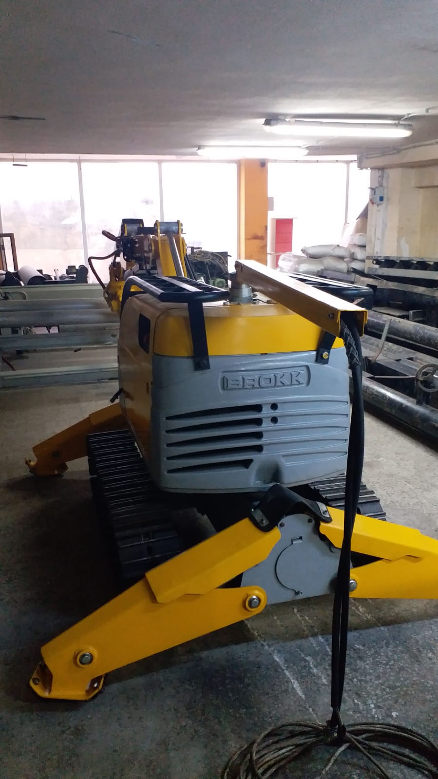 A yellow and gray DEMOLISHING UNIT BROKK 180 REV-F1 with black tracks is parked indoors on a concrete floor near large pipes and industrial materials, with sunlight streaming through the windows in the background.