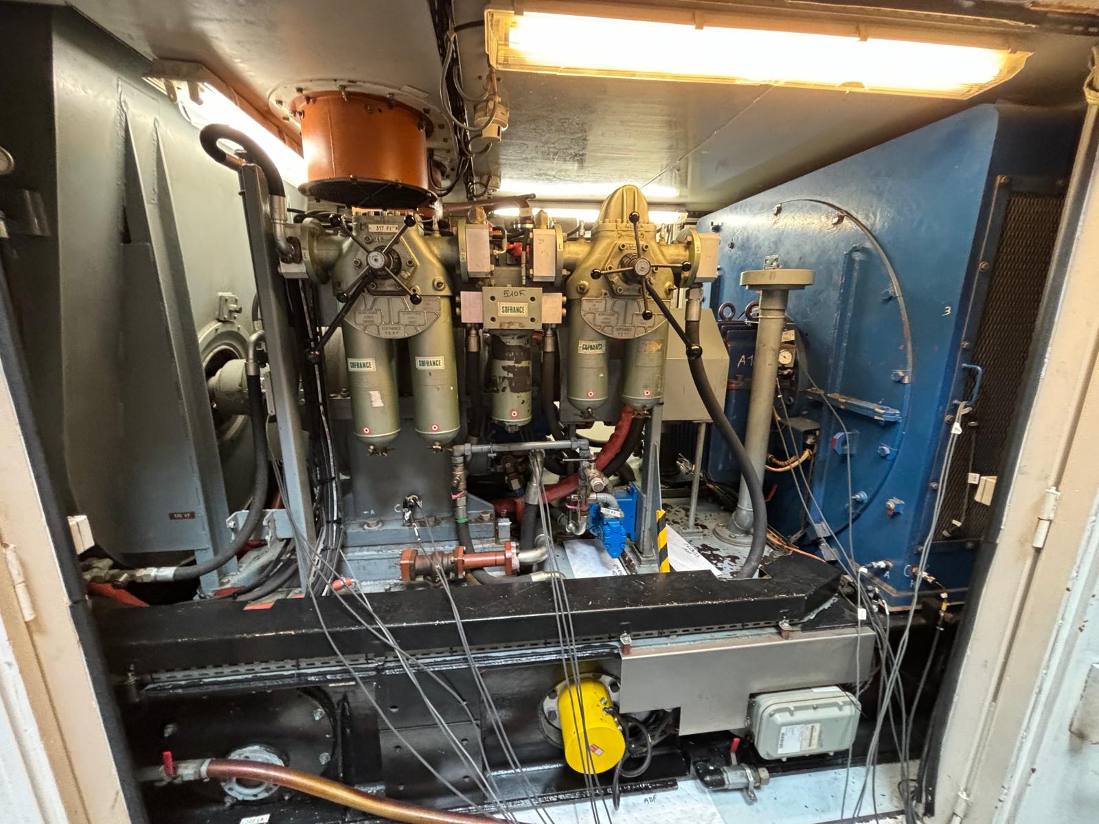 A view inside an engine room with various machinery, pipes, and wires. Large metal components and filters are mounted on the back wall, with a blue panel on the right and a fluorescent light overhead.