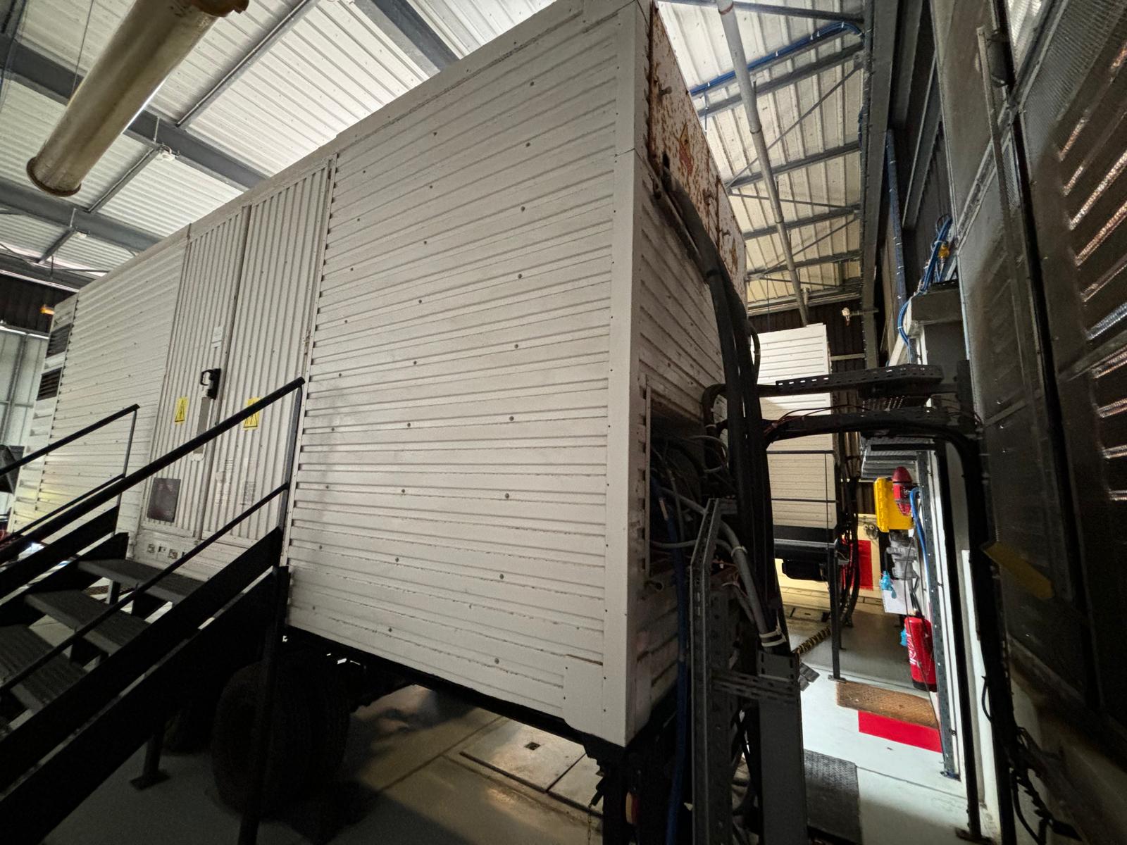 A large white industrial trailer with ribbed metal siding is parked indoors. Stairs lead up to its door, and surrounding equipment is visible in the dimly lit warehouse space.