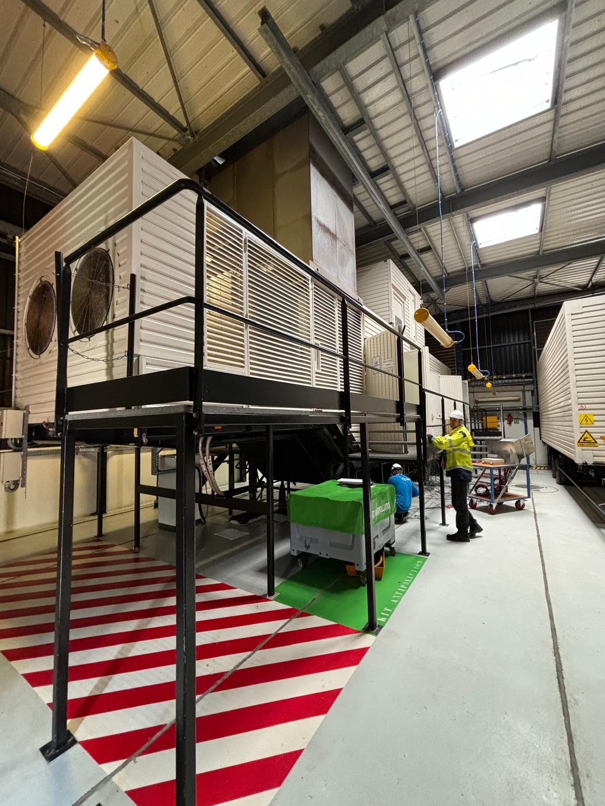 Two workers in safety attire stand beside large industrial HVAC units elevated on metal platforms inside a spacious warehouse with high ceilings and overhead lighting. Striped red and white safety markings are on the floor below.