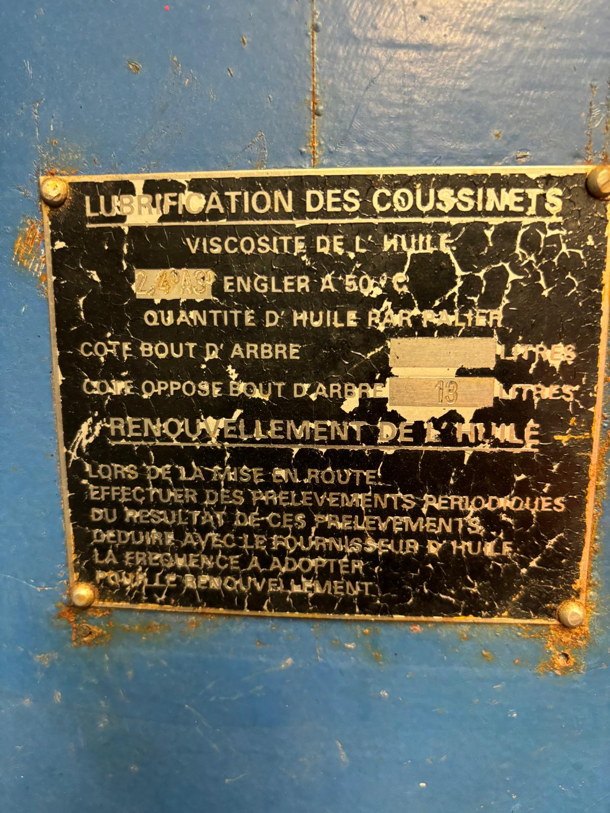 A worn, black and white metal sign in French with text about lubrication instructions for machine bearings. The sign is scratched and partially illegible, mounted on a blue surface.
