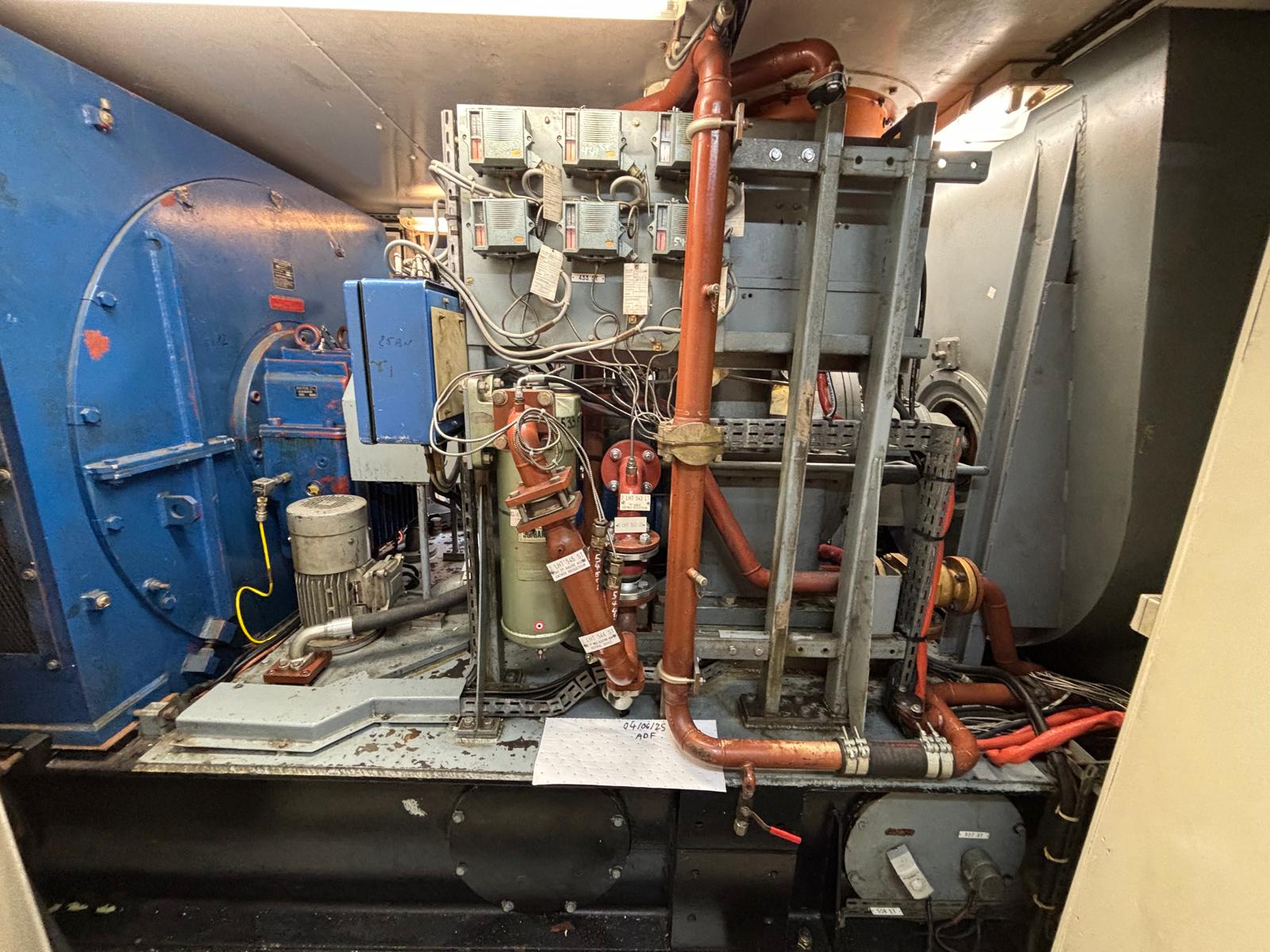 A complex industrial machine with pipes, wires, and gauges is situated in a narrow room with metallic walls. The equipment has a blue cylindrical section on the left and several red and gray pipes and components throughout.
