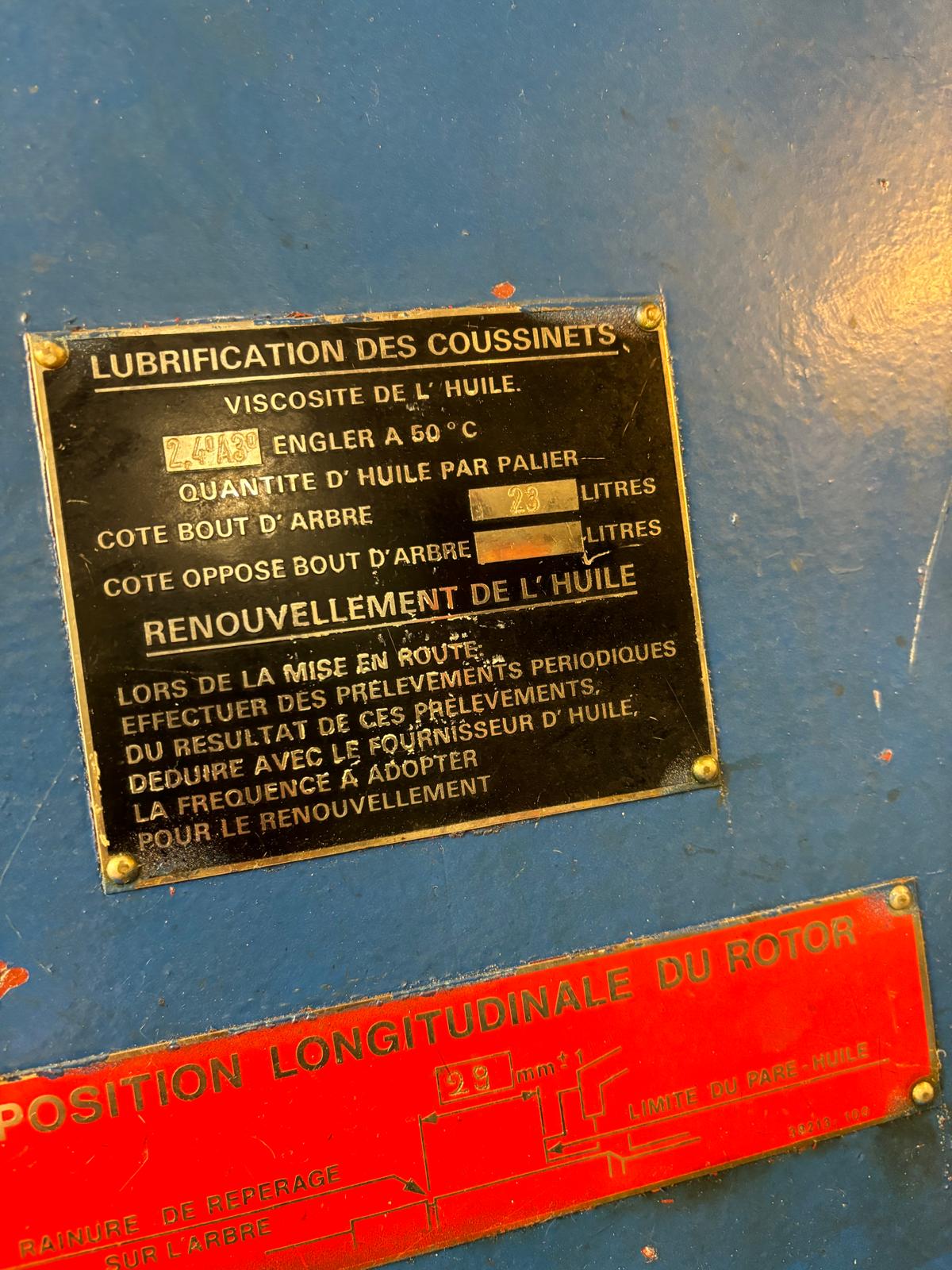 A close-up of a metal equipment panel with French instructions for lubricating bearings, specifying oil viscosity, quantities, and renewal intervals. The panel is slightly worn and mounted on a blue surface with a red label below.