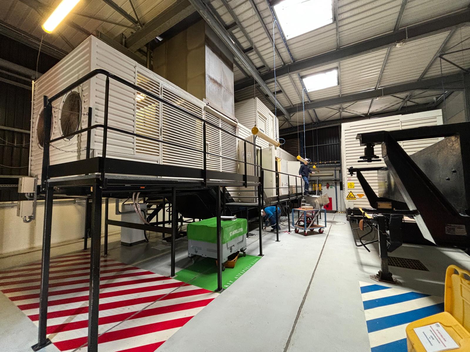 Large industrial equipment inside a spacious facility with metal beams and skylights. A person in safety gear works on a raised platform beside machinery. Colored floor markings indicate safety zones.
