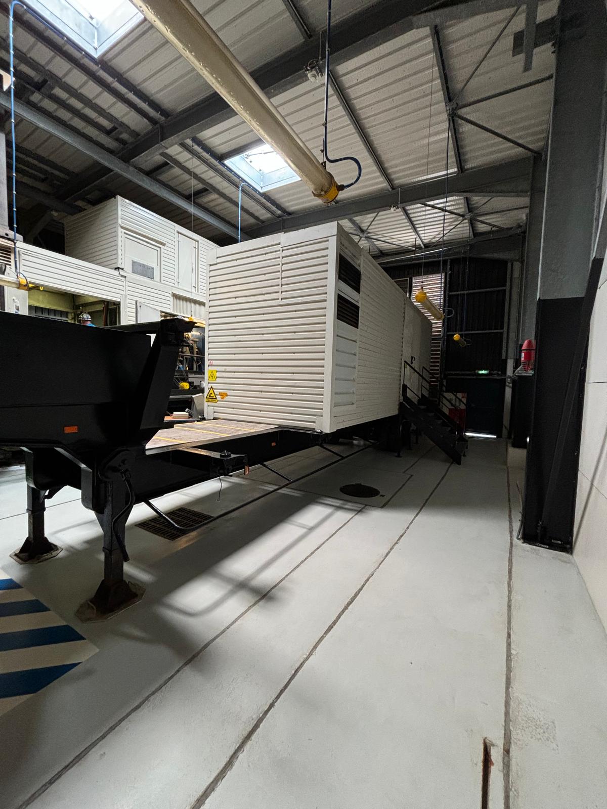 A large white modular unit is positioned on a trailer inside an industrial building, with metal beams, overhead pipes, and a concrete floor visible in the surrounding warehouse space.