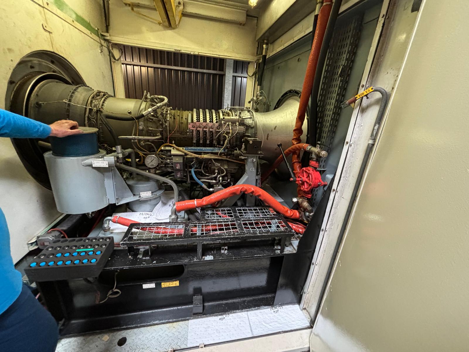 A person in a blue shirt operates or inspects a large industrial machine with numerous pipes, wires, and metal components inside a confined space. An orange cable and red valve are visible in the foreground.