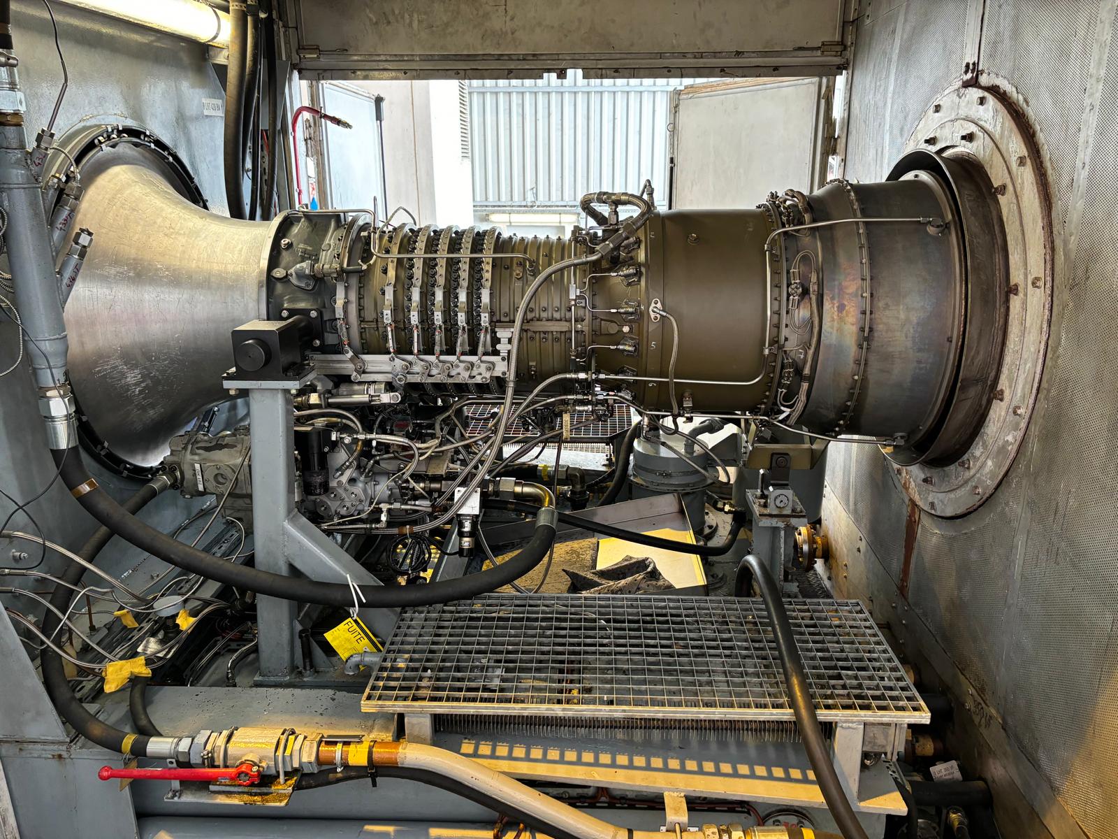 A close-up view of an industrial gas turbine engine mounted inside a metallic testing facility, surrounded by cables, pipes, and metal grates, with a vented background wall.