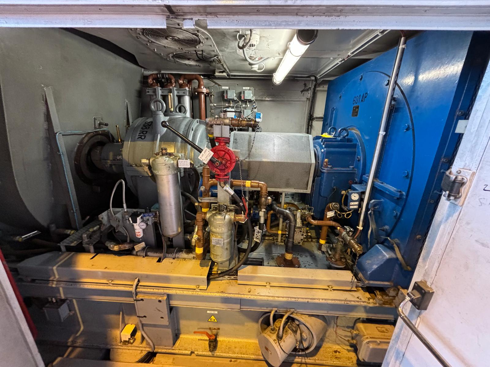 A large industrial machine with blue, silver, and metallic components, pipes, and gauges inside an enclosed area, illuminated by overhead lighting. Various cables and mechanical parts are visible.