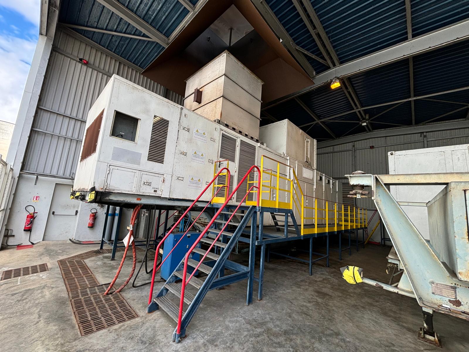 Large industrial air handling units are elevated on a metal platform with yellow railings and accessed by blue stairs, inside a spacious warehouse with a high ceiling and fire extinguishers on the left wall.