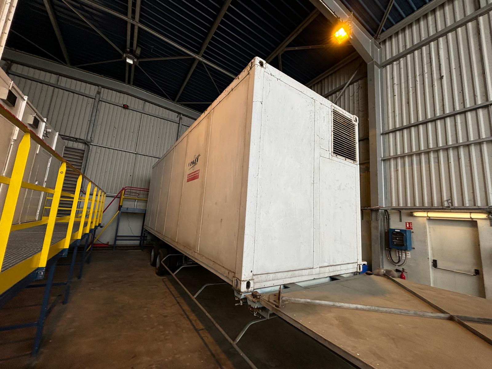 A large white refrigerated trailer is parked inside an industrial warehouse, positioned next to a yellow railing and metal stairs, with overhead lights illuminating the space.