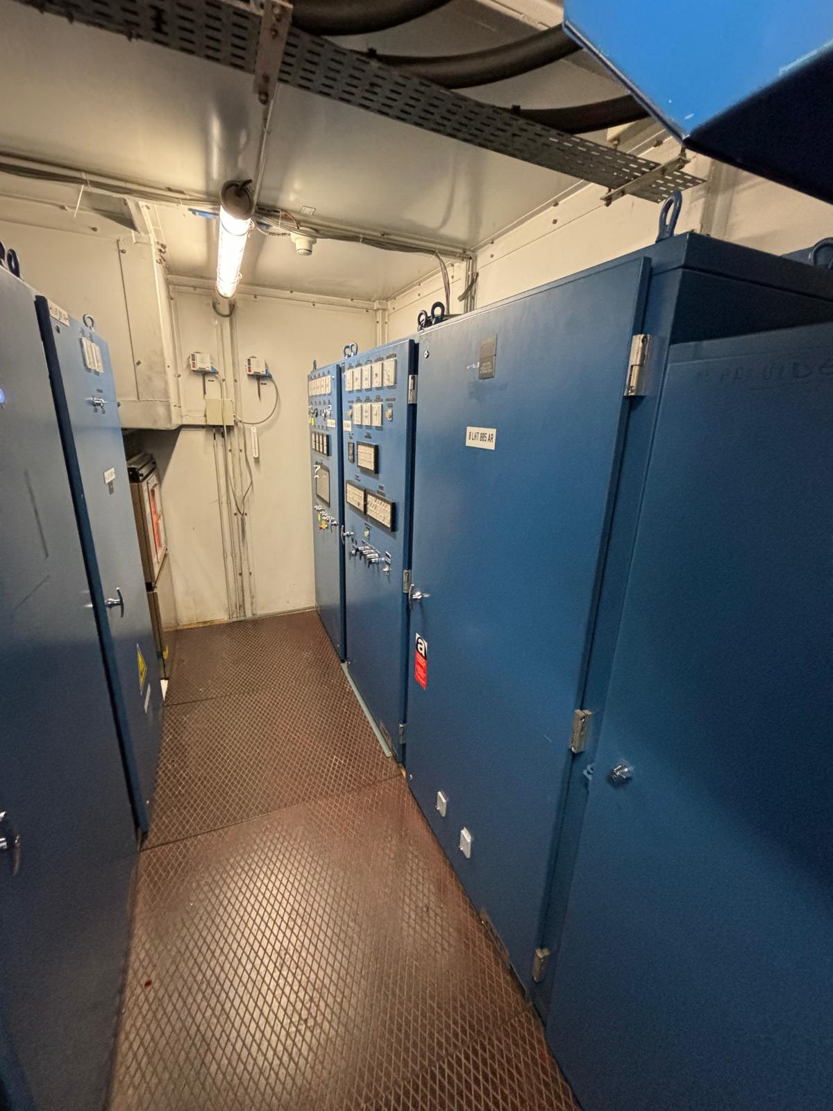 A narrow industrial room with blue electrical control panels on both sides, featuring dials, switches, and meters; overhead fluorescent lighting and metal flooring are visible.