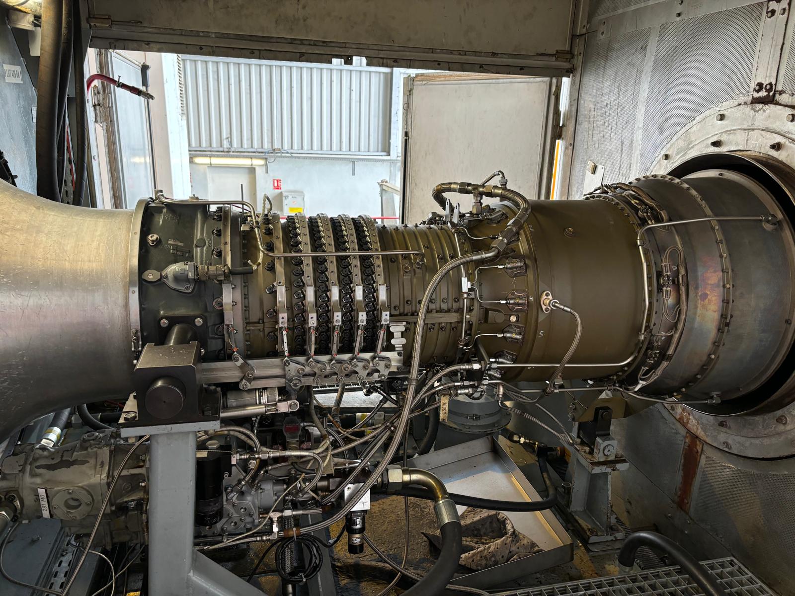 A close-up view of a large industrial gas turbine engine mounted on a test stand inside a facility, with various cables, pipes, and metal components visible around the cylindrical engine.
