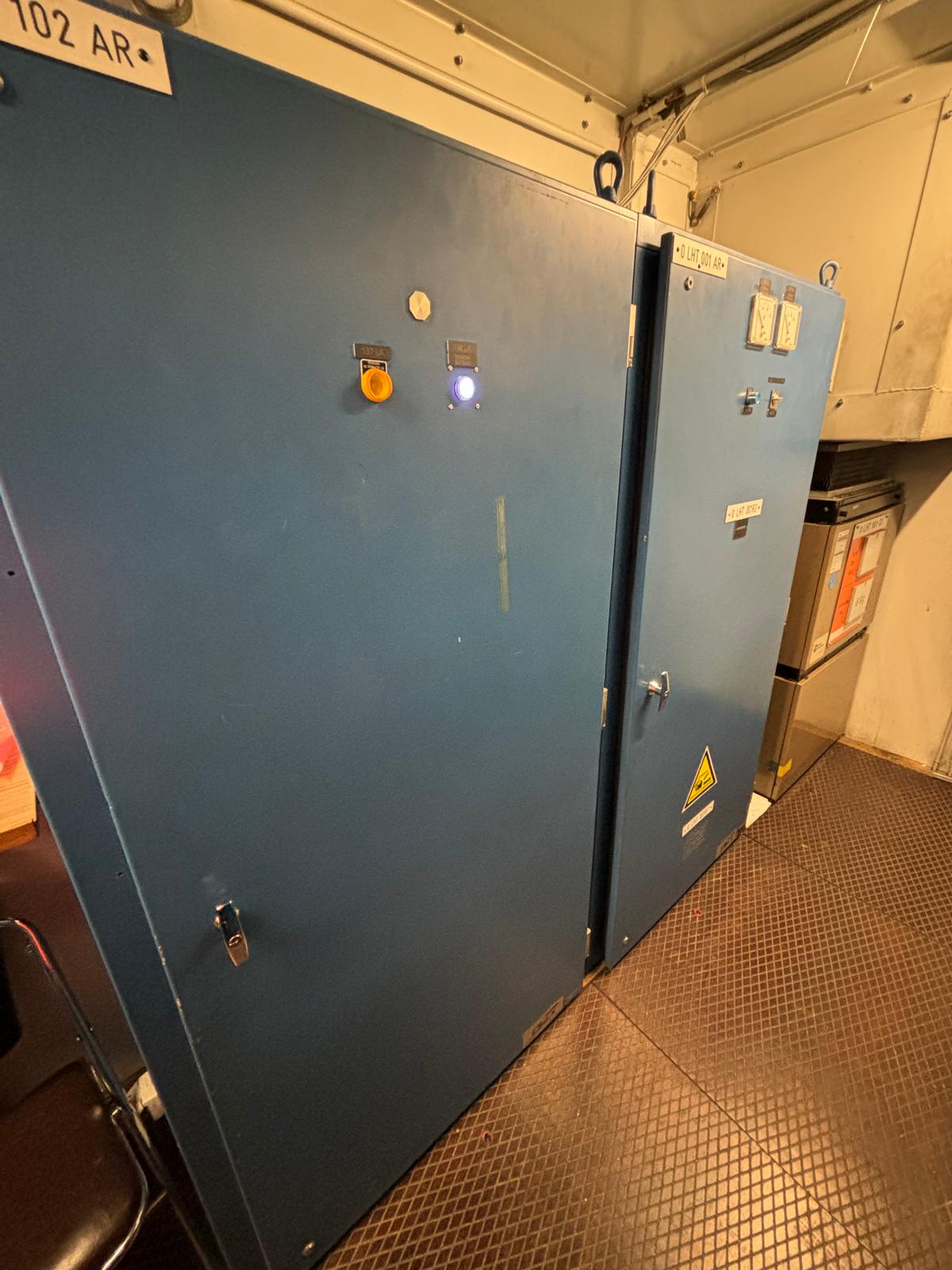 Large blue industrial electrical cabinets with control buttons, indicator lights, and warning labels are placed against a wall in a room with metal flooring. The cabinets have closed doors and labels with numbers.