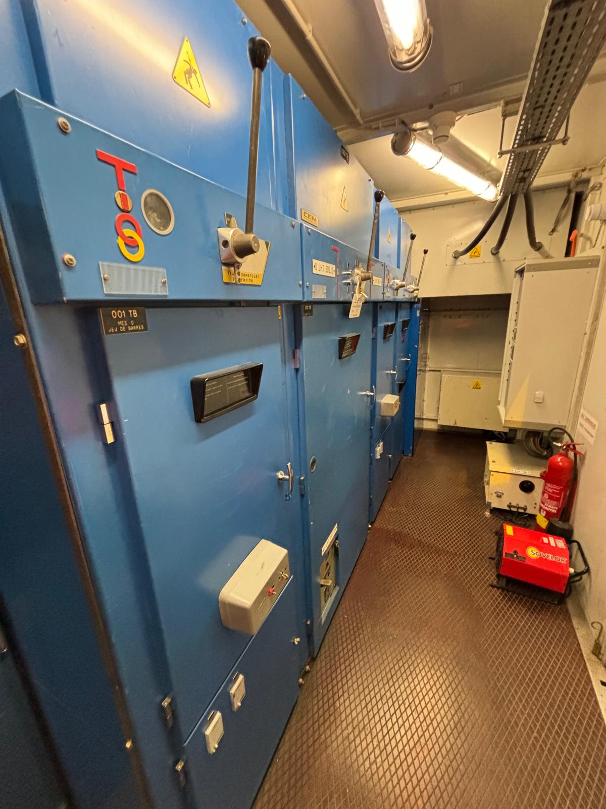 A narrow electrical control room with large blue metal cabinets along one wall, labeled with warning signs and switches. A fire extinguisher, open panel, and red equipment are visible on the right side of the room.
