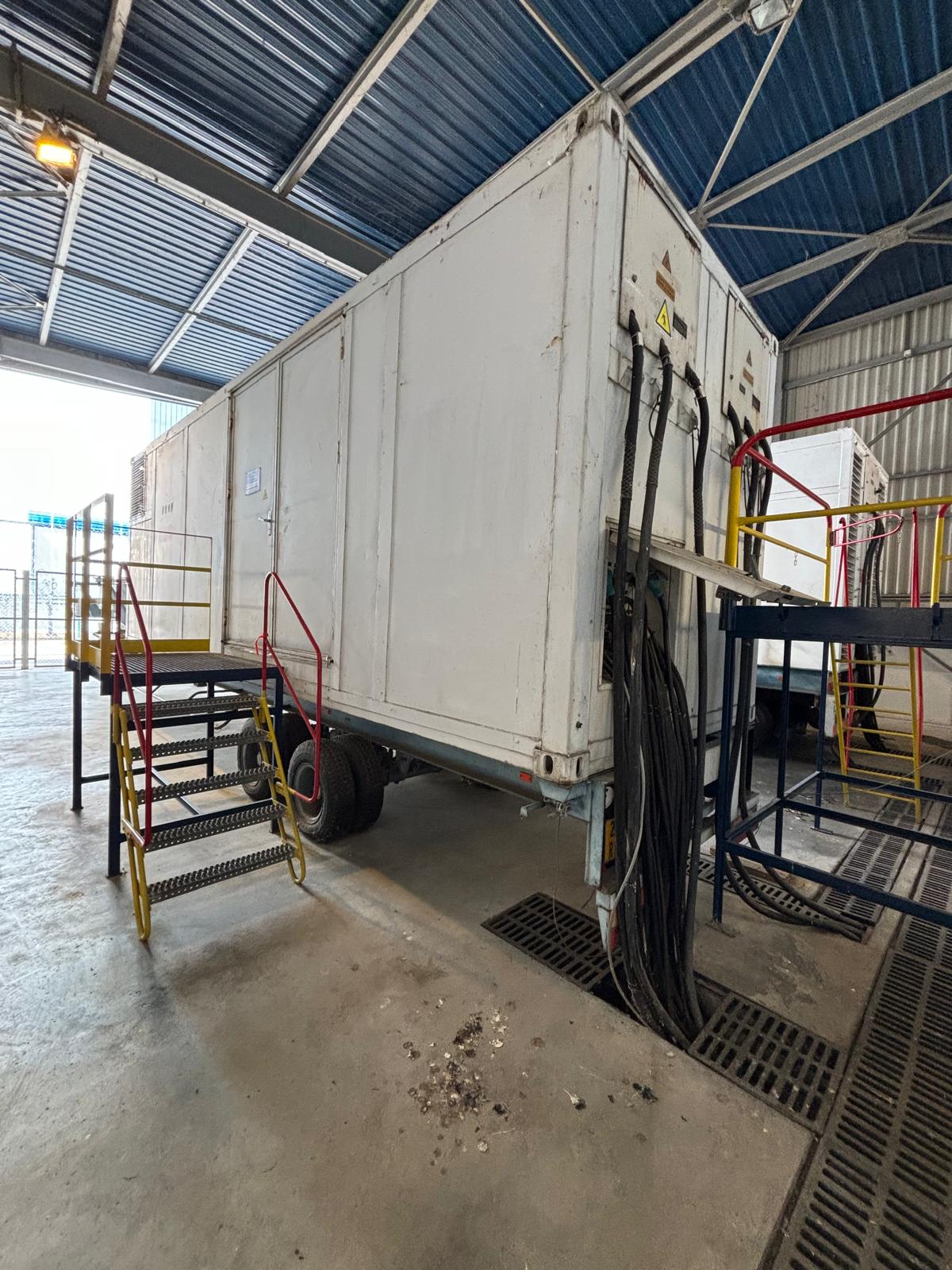 A large white trailer with cables attached is parked indoors under a metal roof. Two staircases with yellow and red railings lead to platforms on either side of the trailer. The area is industrial and well-lit.