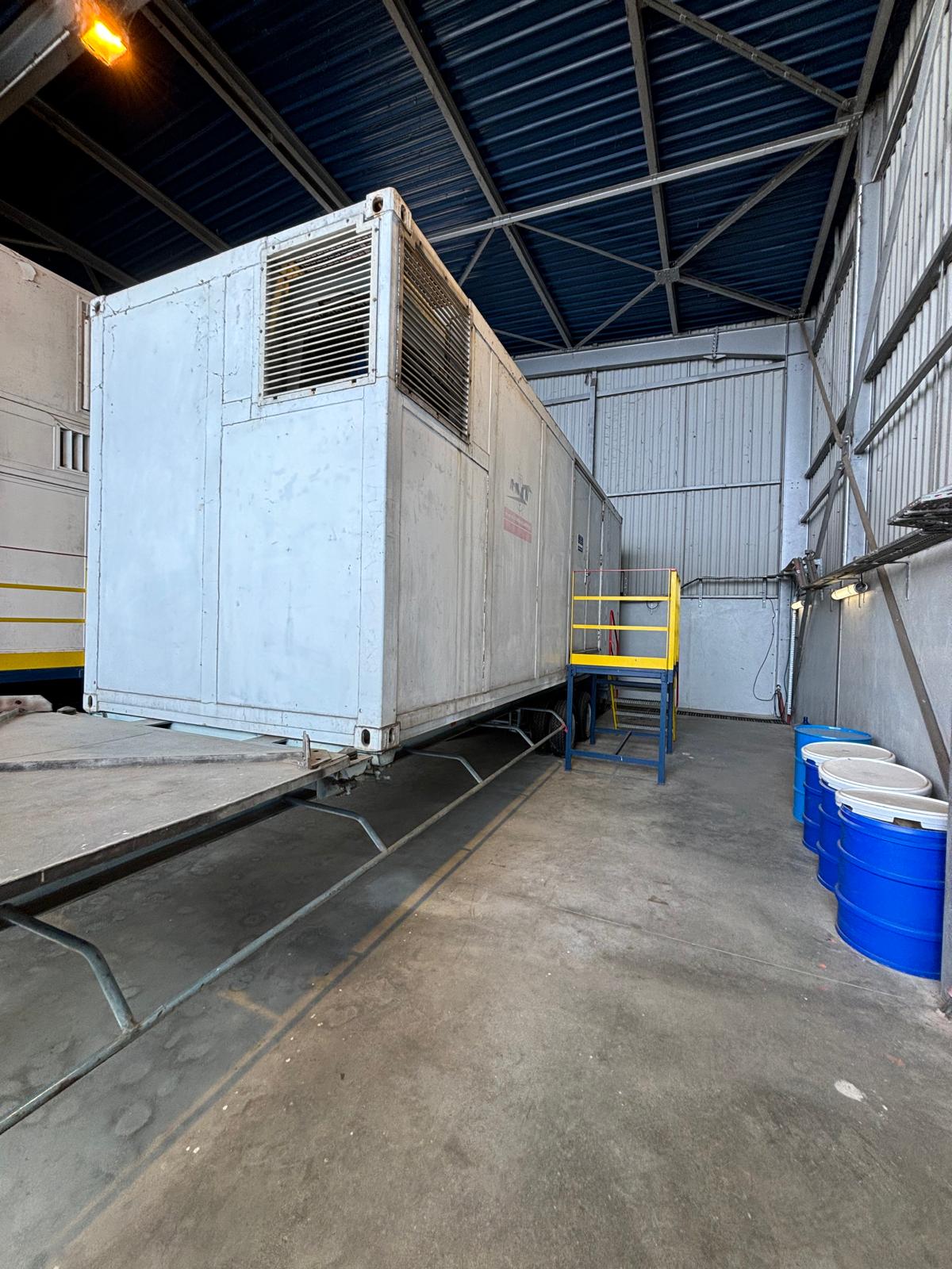 A large white industrial container with vents is elevated on a platform inside a metal warehouse. Blue barrels are lined up on the right, and yellow railings and stairs lead to the platform. The floor is concrete.