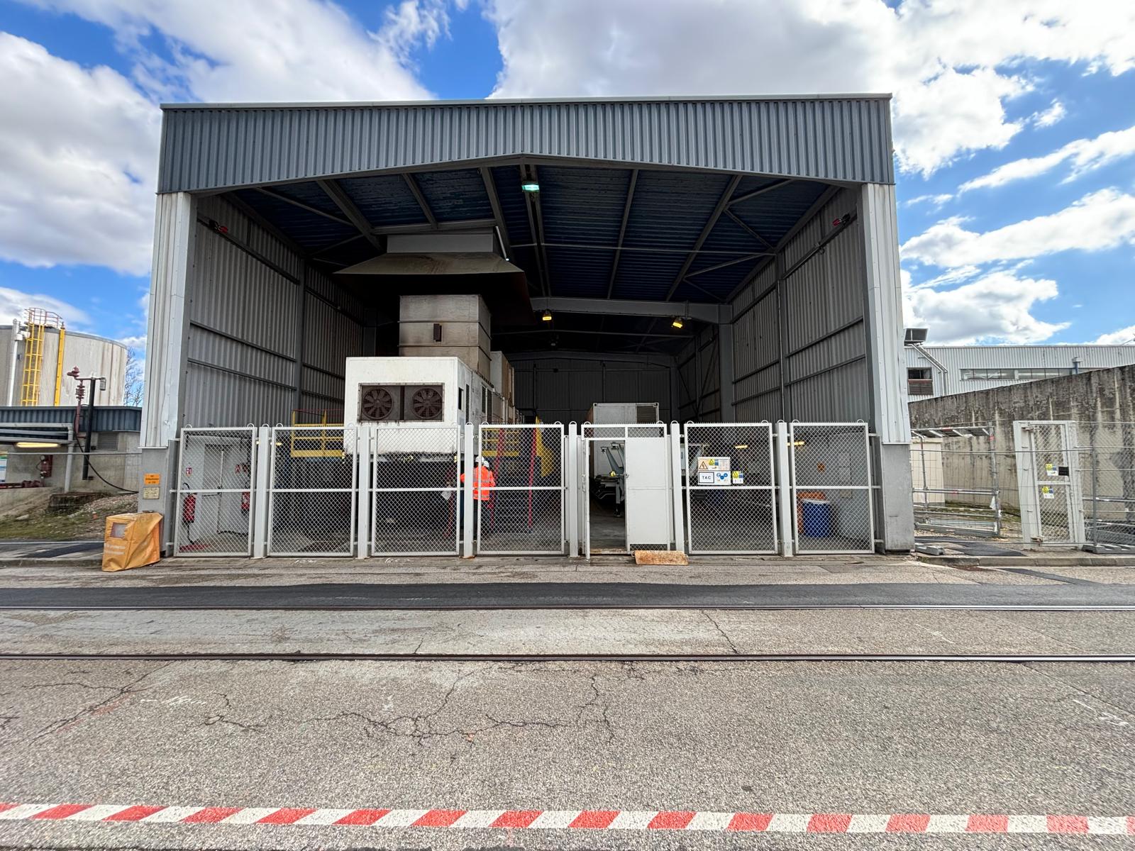 An industrial facility with a large open-front metal building, fenced area, machinery inside, and a partly cloudy sky above. The ground has railroad tracks and a red-white striped safety marking in the foreground.