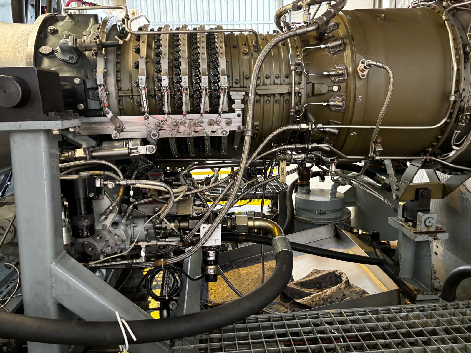 Close-up view of a complex jet engine, showing metal pipes, wires, and mechanical components assembled on a support structure inside an industrial setting.