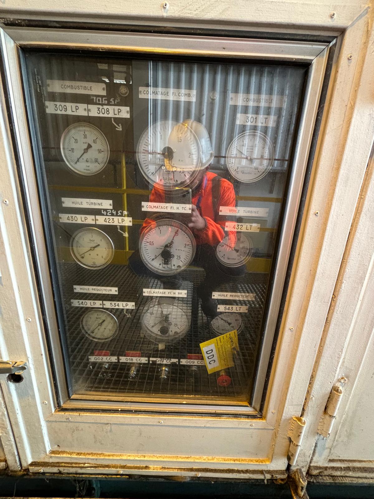 A person in a red safety vest and helmet is reflected in a glass panel displaying multiple analog pressure gauges, each labeled with numbers and descriptions. The setting appears industrial, with visible pipes and metal structures.