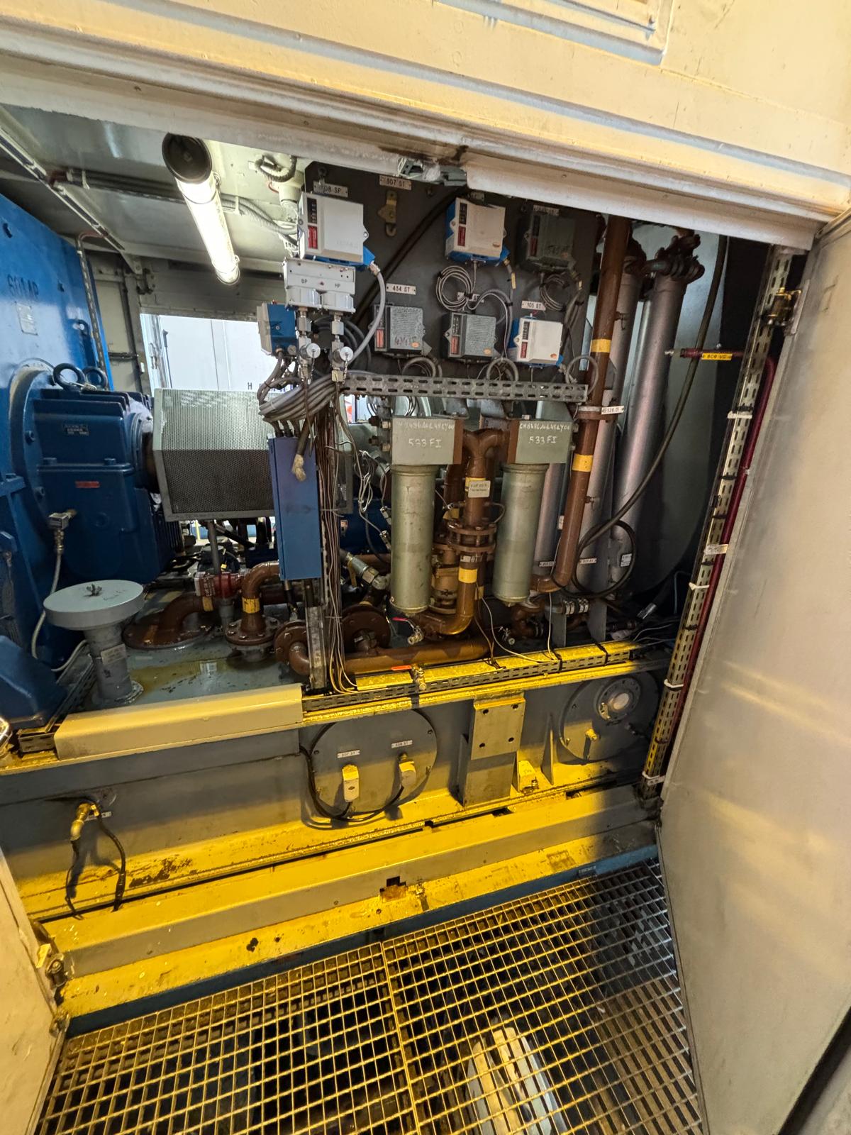 An industrial machine room with pipes, filters, gauges, and electrical boxes, viewed through an open door. The floor is grated metal, and yellow guardrails are present around the machinery.
