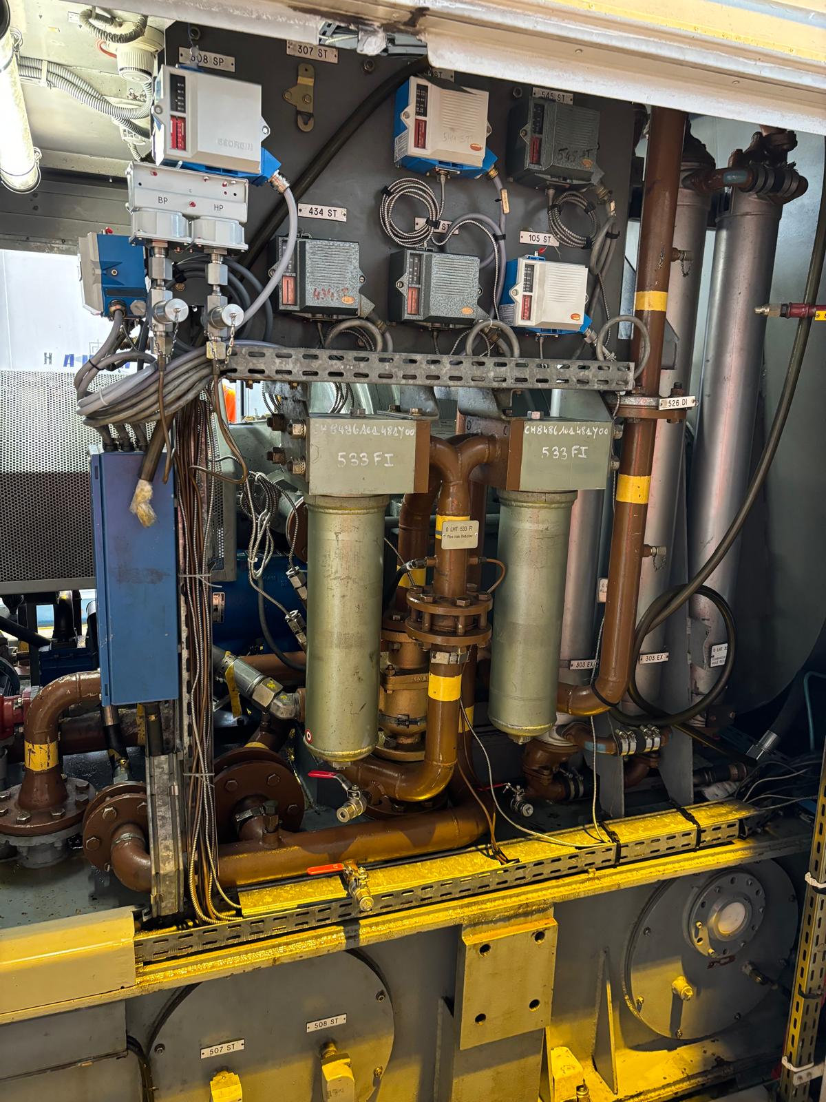 Close-up of industrial machinery with pipes, wires, and control boxes, featuring two large vertical cylinders labeled “GENERAL FILTRATION 533 FE,” surrounded by various gauges and connected metal components.