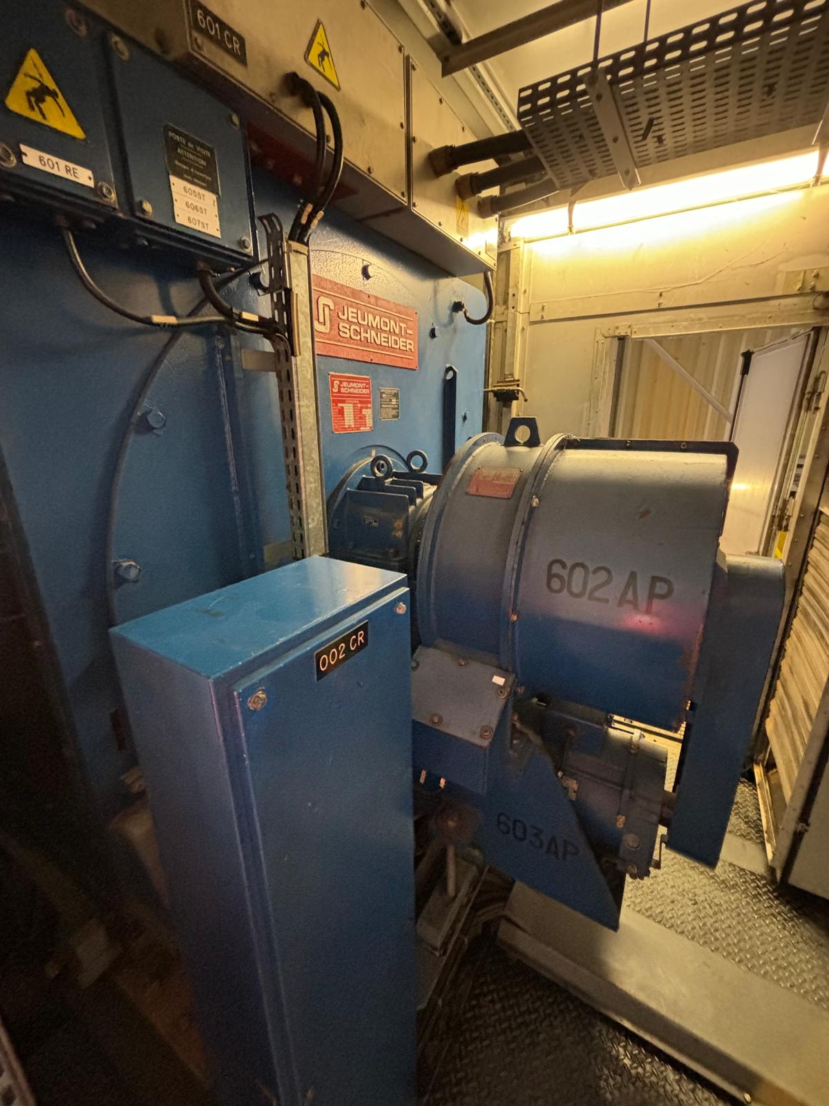 A close-up view of industrial blue electrical equipment labeled Jeumont Schneider and marked 602AP inside a metal enclosure, with warning signs and wired components in a confined, lit space.