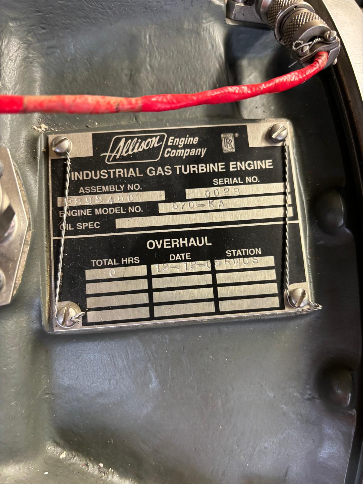 Metal nameplate on industrial gas turbine engine showing assembly, serial, and model numbers, oil specification, and overhaul section for total hours, date, and status. Red cable and nearby wires are visible.