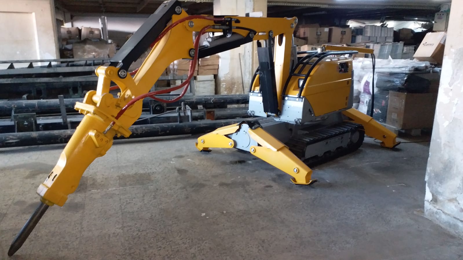 A compact, yellow DEMOLISHING UNIT BROKK 180 REV-F1 with a hydraulic arm and breaker is positioned indoors on a concrete floor, surrounded by industrial materials and equipment.