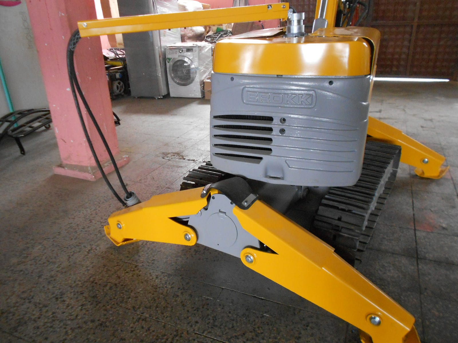 A close-up of a DEMOLISHING UNIT BROKK 180 REV-F1 in yellow and gray, with treads, indoors on a tiled floor, surrounded by appliances and other objects in the background.