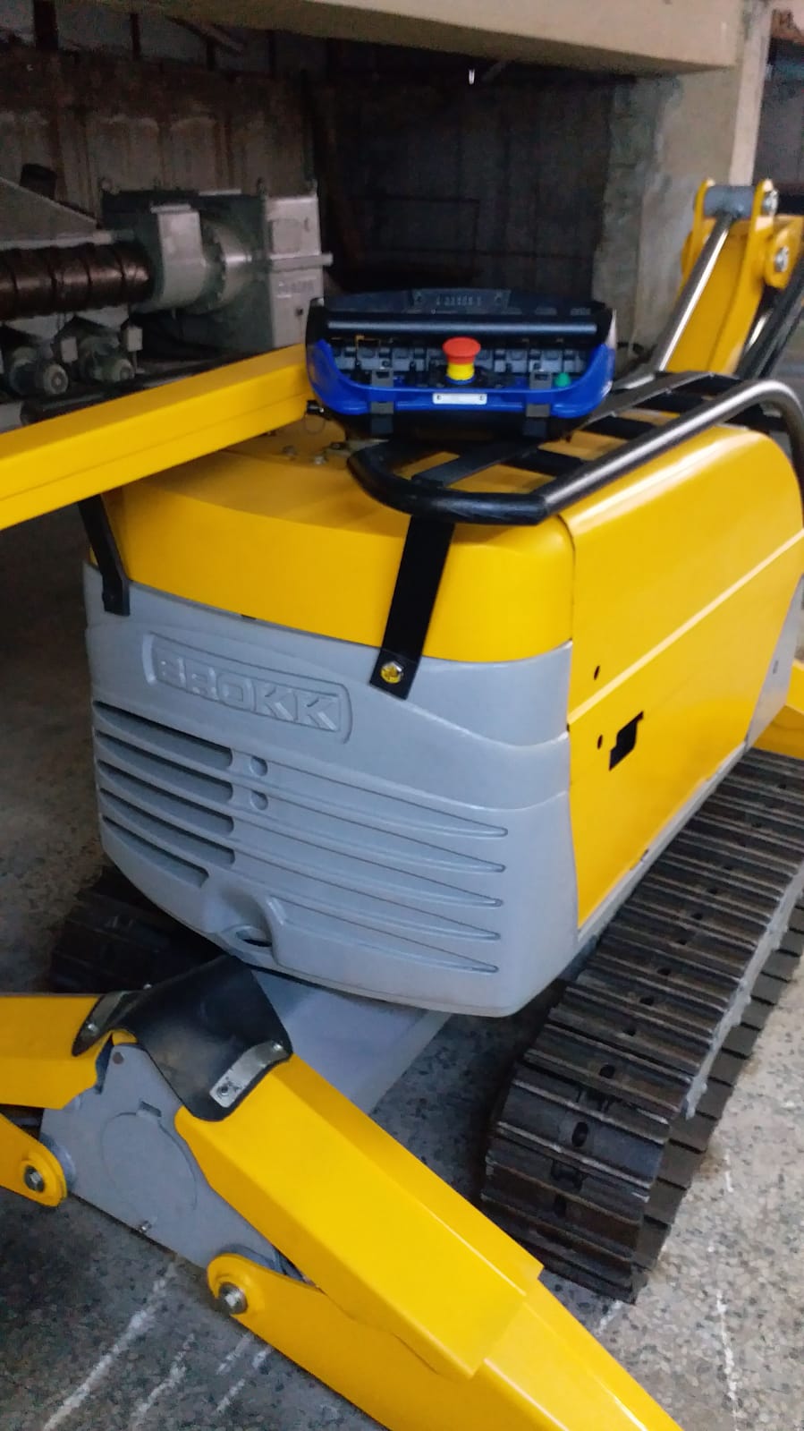 A DEMOLISHING UNIT BROKK 180 REV-F1 in yellow and gray with rubber tracks is parked indoors; a blue remote control with buttons and a red emergency stop sits atop the machine.