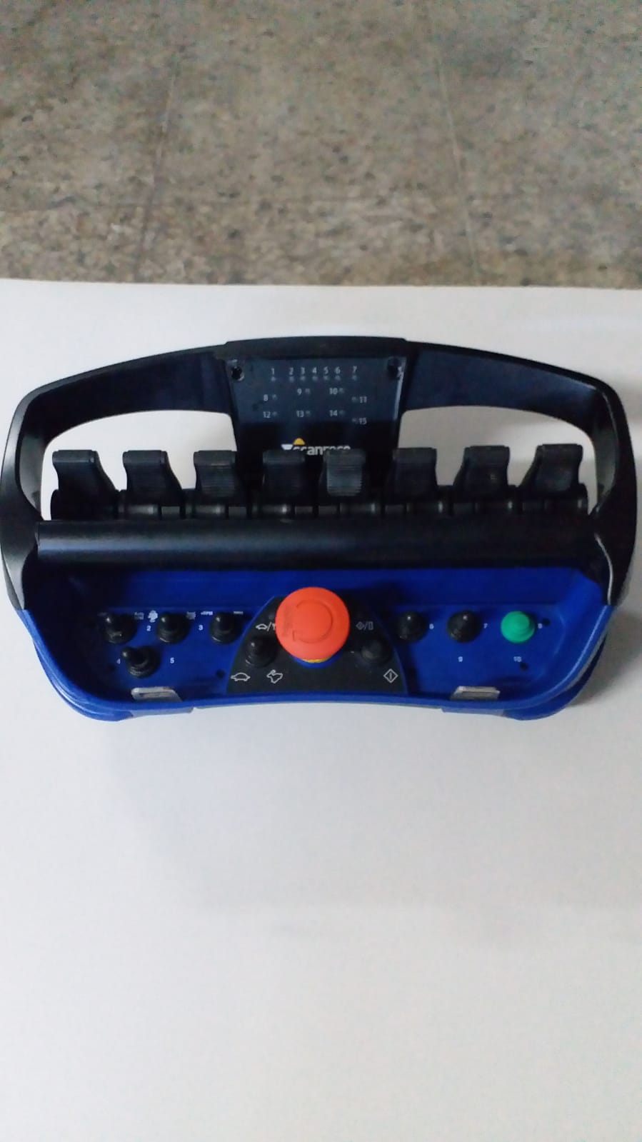 A blue and black handheld remote control for the DEMOLISHING UNIT BROKK 180 REV-F1, featuring several levers, buttons, and a large red emergency stop button, is placed on a white surface.