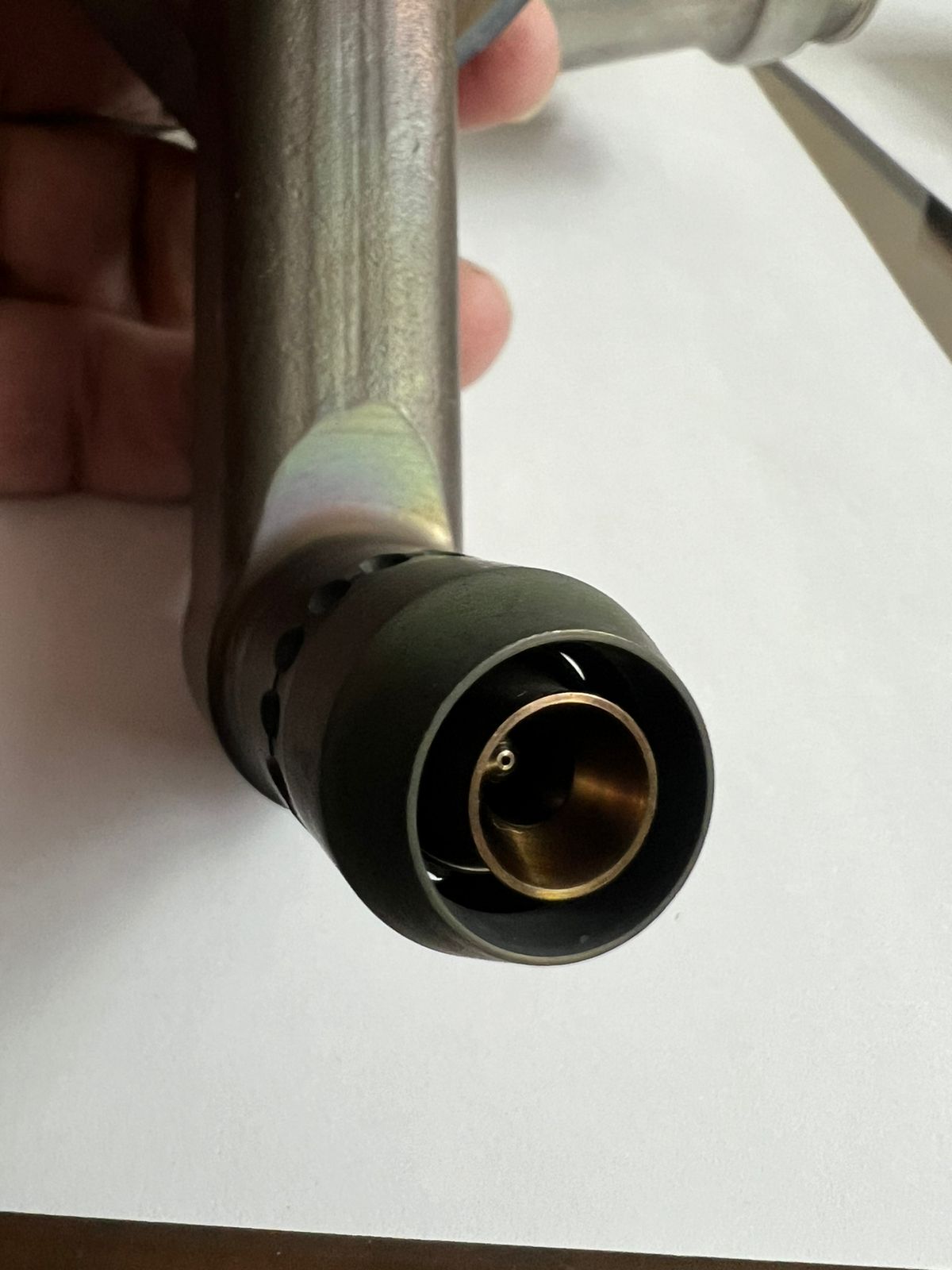A close-up of a hand holding a metallic device with a cylindrical, brass-colored nozzle at the end, possibly part of a mechanical or plumbing component, set against a white background.