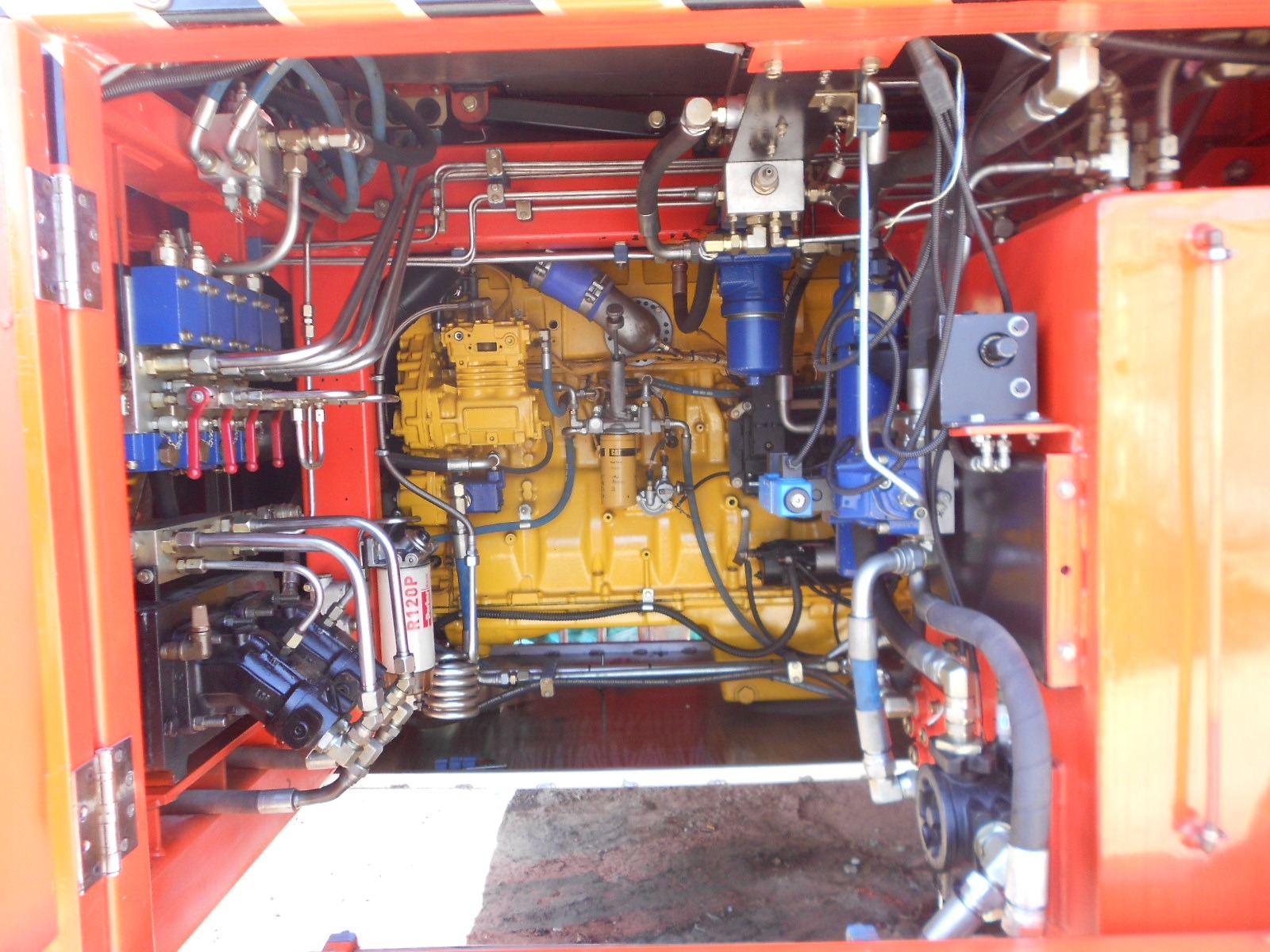 Close-up of a complex hydraulic and mechanical system inside a machine, featuring yellow, red, and blue components, various pipes, hoses, metal parts, and electrical wiring.