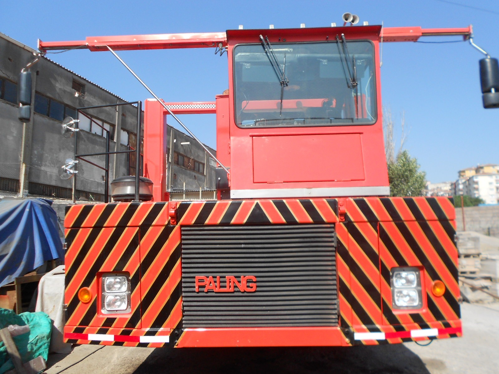 Front view of a large red industrial vehicle with black and yellow diagonal safety stripes. The vehicle has PALFINGER written on the front grill and is parked outdoors near buildings on a sunny day.