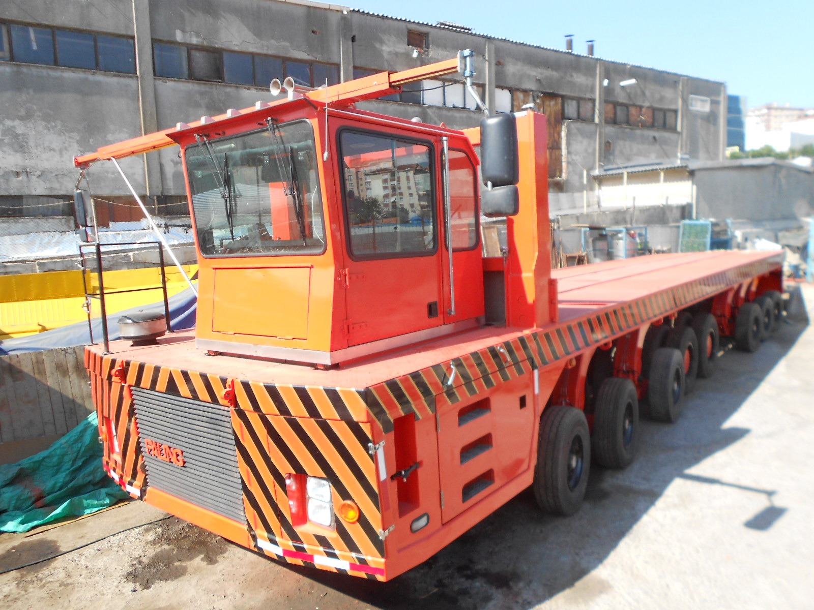 An industrial, bright orange multi-axle heavy transport vehicle with a large elevated operator cab and striped warning markings, parked in a construction yard near a weathered concrete building.