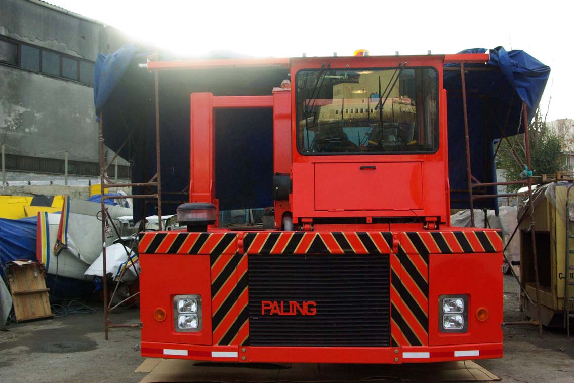 A large AVAILABLE PALING HEAVY TRANSPORTER stands in a cluttered outdoor iron mill, black and yellow hazard stripes on the front grill marked "PALING," surrounded by blue tarps and scattered equipment.