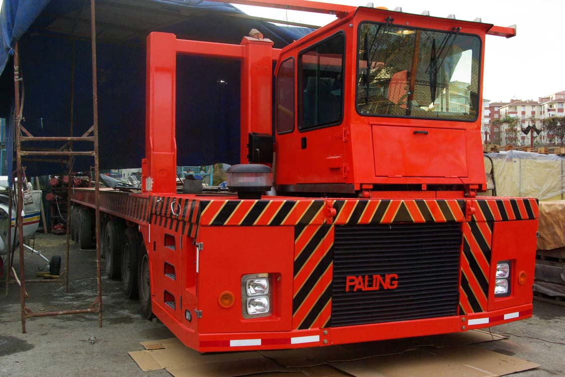 The AVAILABLE PALING HEAVY TRANSPORTER, a large red heavy-duty truck with black and yellow safety stripes, is parked outdoors near construction materials and an Iron Mill building. "Paling" is displayed on the front.