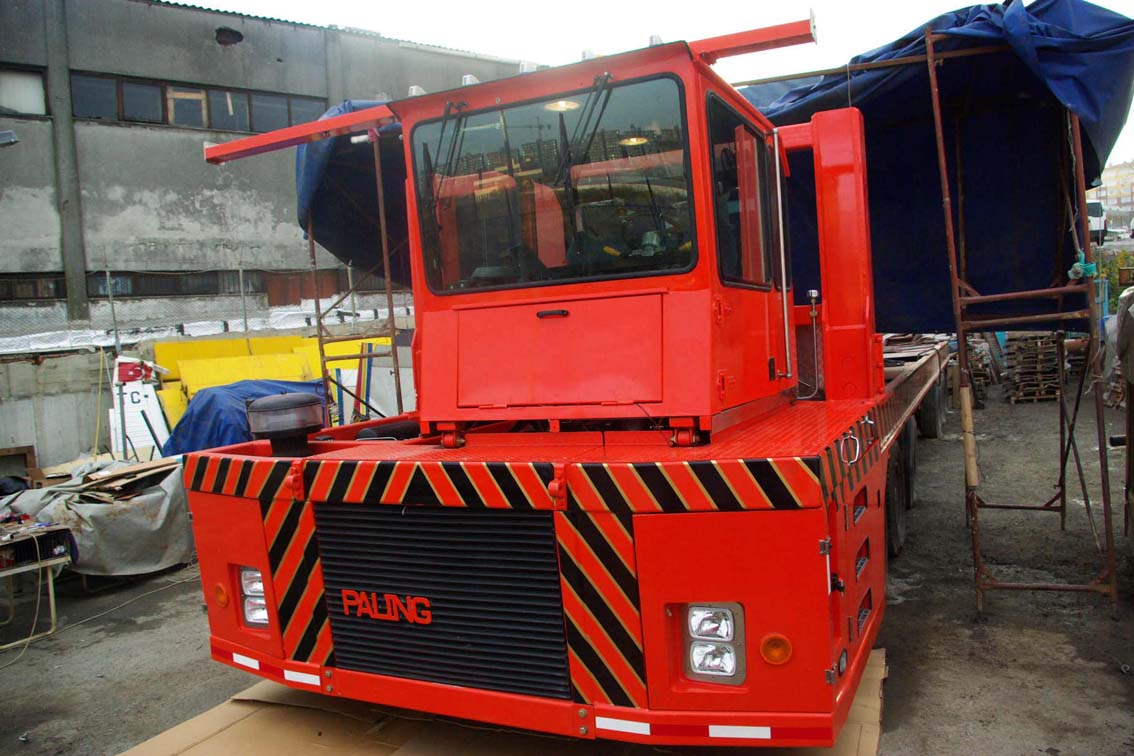 A bright red AVAILABLE PALING HEAVY TRANSPORTER with black and yellow hazard stripes on the front is parked outdoors near an iron mill, surrounded by buildings and covered materials. "PALING" is visible on its front grille.