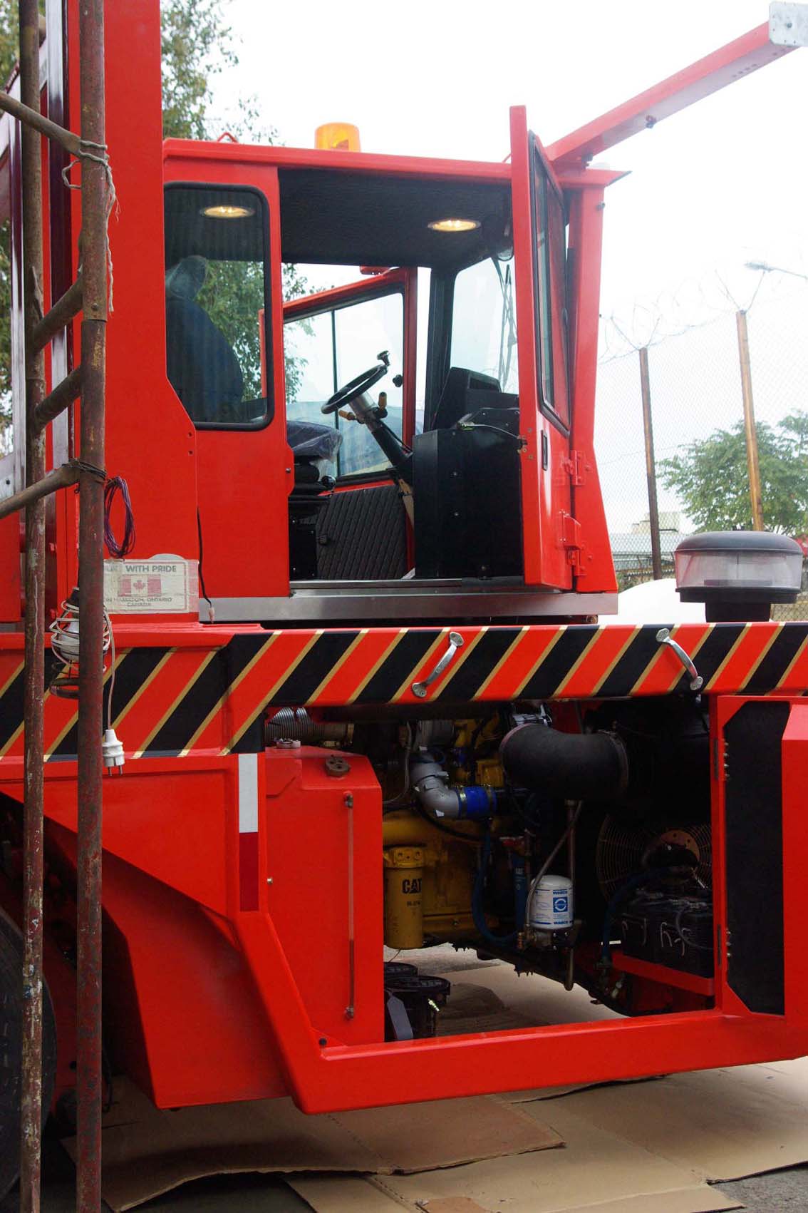 A close-up of the AVAILABLE PALING HEAVY TRANSPORTER, a bright red industrial vehicle with black and yellow hazard stripes, highlighting its driver’s cab and engine at the iron mill, with a metal fence and trees in the background.