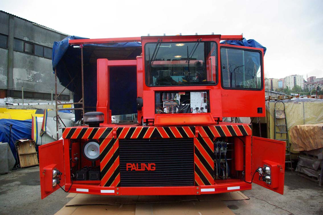 A large, red AVAILABLE PALING HEAVY TRANSPORTER with its panels open to reveal mechanical parts stands outdoors near iron mill buildings and assorted equipment, with "PALING" displayed on the front.