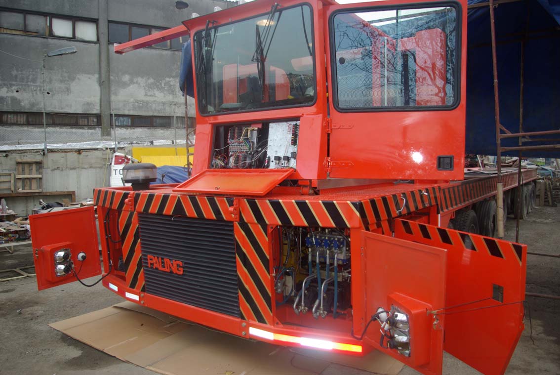 The AVAILABLE PALING HEAVY TRANSPORTER, a large bright orange vehicle with red and black stripes, has its front panels open to show internal mechanical and electrical parts. Its elevated cab features glass windows in an iron mill setting.