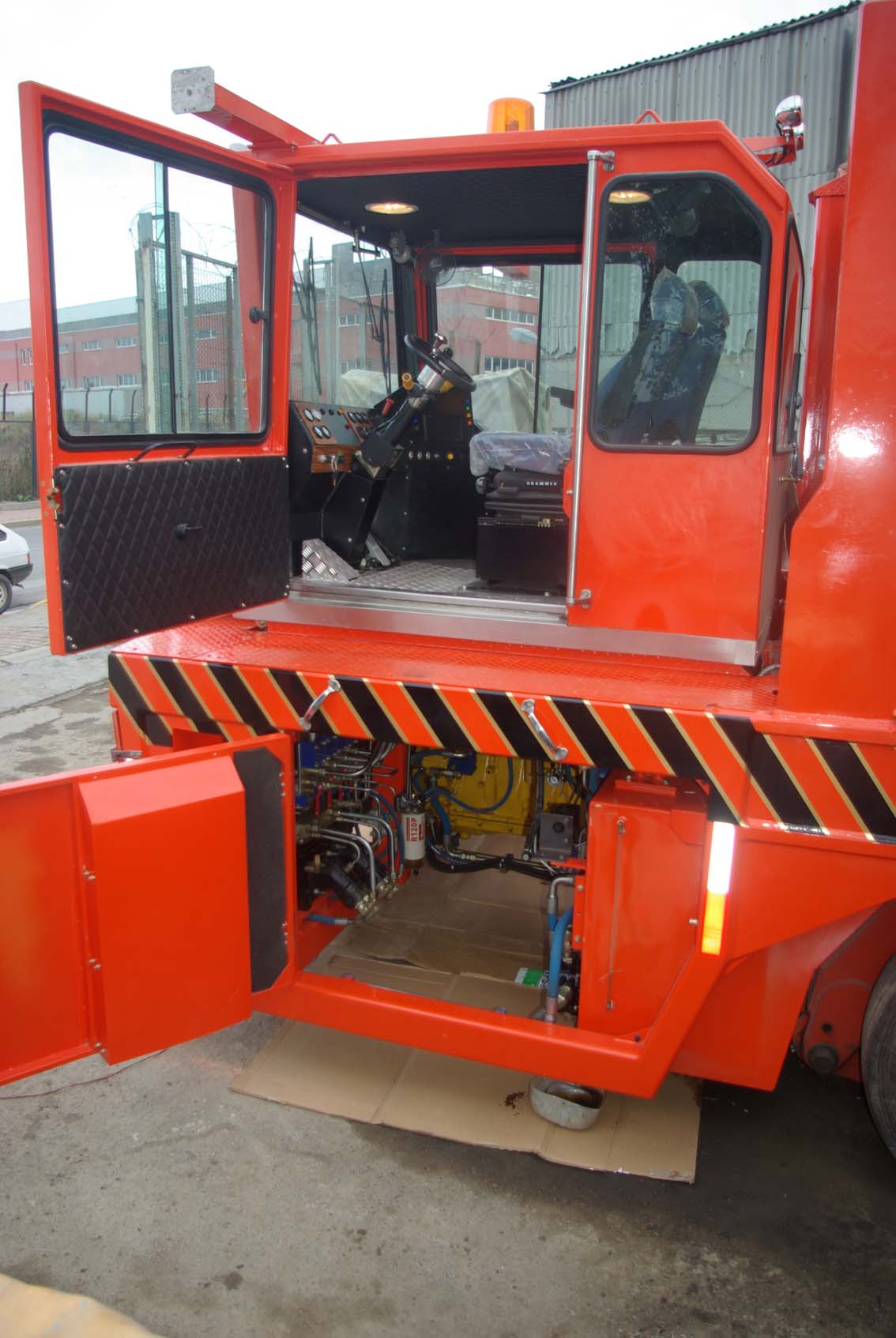 The AVAILABLE PALING HEAVY TRANSPORTER, a bright orange utility vehicle with its cab door and rear compartment open to show control panels and mechanical parts, is parked outdoors near an iron mill and industrial fencing.