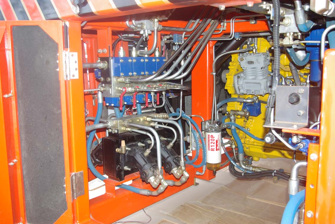 Close-up of an AVAILABLE PALING HEAVY TRANSPORTER, showcasing exposed wiring, pipes, mechanical parts, hydraulic lines, blue and yellow components in a bright orange frame, and a white Fleetguard fuel filter.
